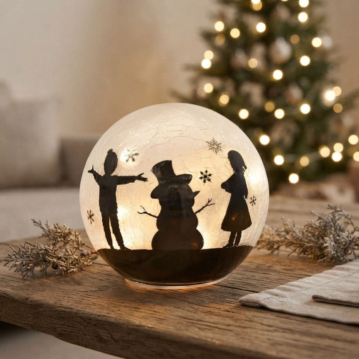 A glowing round glass ornament or decorative globe with black silhouettes of children, a snowman, and snowflakes, placed on a wooden table in a cozy indoor setting with a blurred Christmas tree in the background.