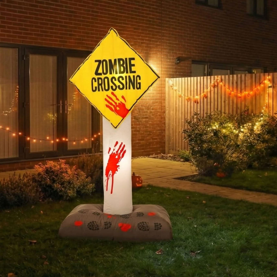 A Halloween yard decoration shaped like a 'Zombie Crossing' sign on a white post, set on a gray weighted base in a backyard at night. The sign is yellow with black text and a red handprint, and the post has a red handprint near the bottom.