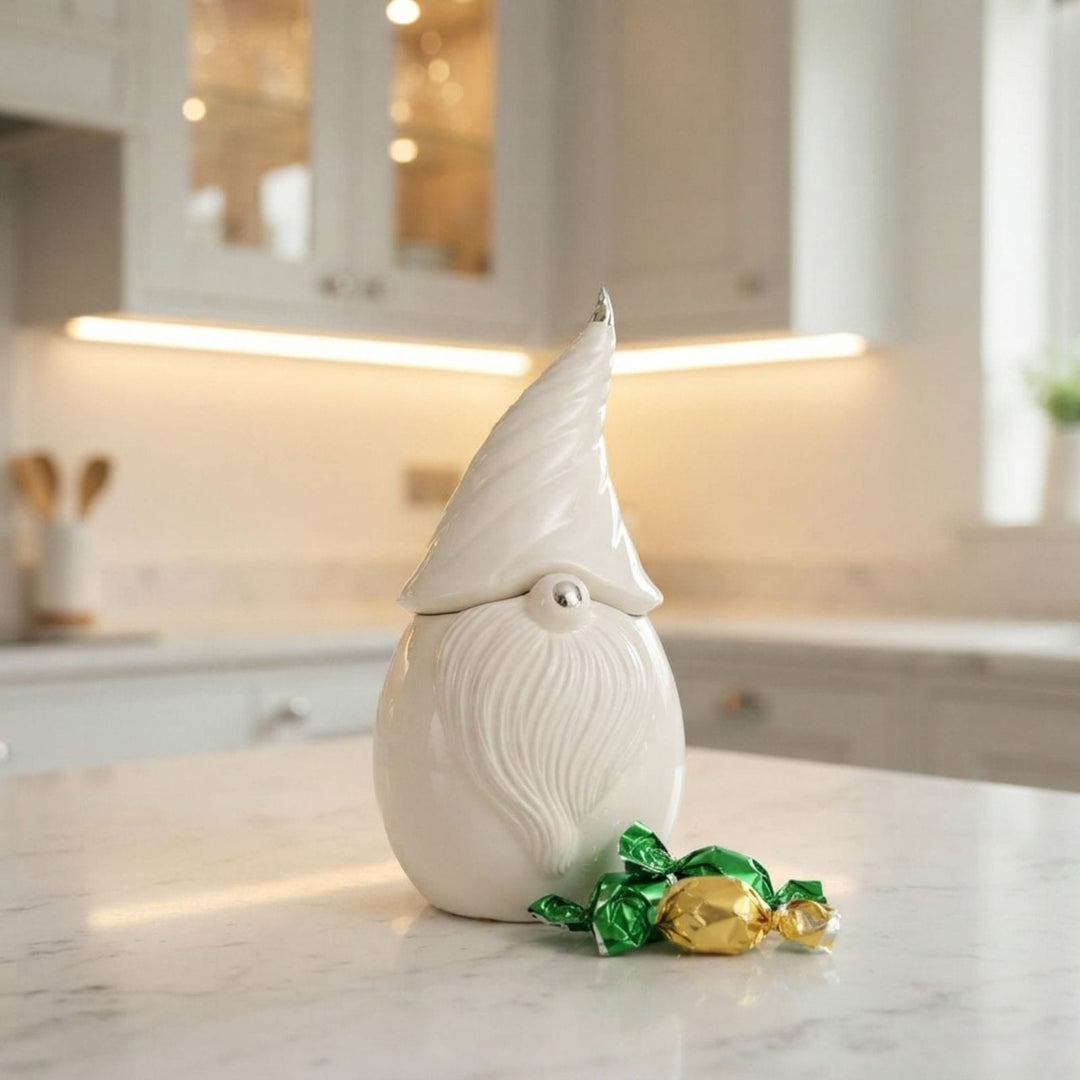 A white ceramic gnome figurine with a textured beard and pointed hat sits on a marble kitchen counter, with foil-wrapped chocolates in green and gold in front of it.