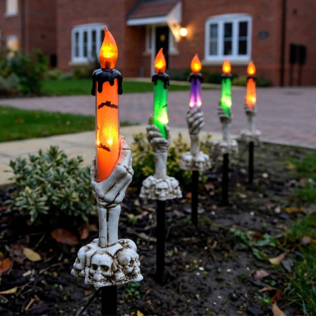 Halloween yard decorations featuring skeleton hands holding glowing LED candles with black wax details, arranged in a row along a garden path in front of a brick house.