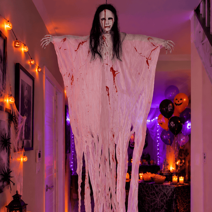 A Halloween hanging ghost decoration with a pale pink tattered sheet body, black hair, and red blood splatter, suspended in a decorated home hallway with string lights, candles, and balloons.