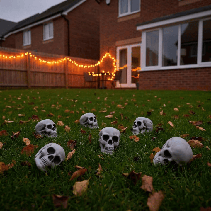 Six white skull decorations arranged on a green lawn scattered with autumn leaves, set in a backyard with warm string lights and a brick house in the background.