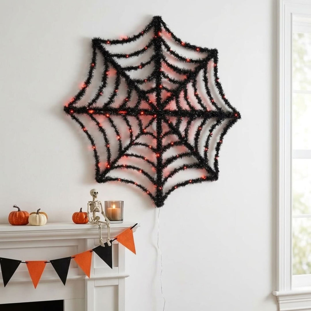 A large black spiderweb wall decoration with warm white string lights, displayed on a white wall above a decorated mantel with pumpkins and a skeleton.
