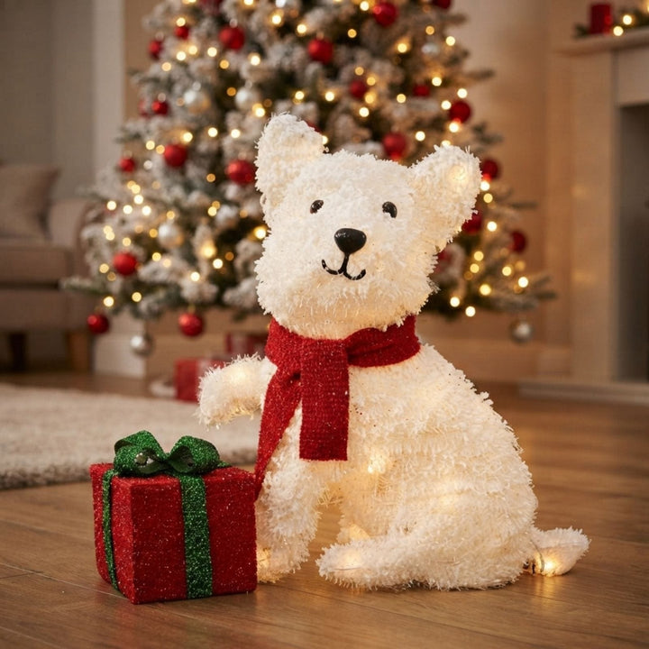 A fluffy white dog Christmas decoration with a red scarf sits on a wooden floor beside a small red gift box with a green ribbon, with a lit Christmas tree in the background.