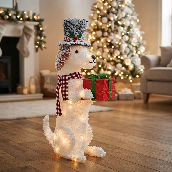 A fluffy white Christmas dog decoration wearing a gray top hat with red berries and a red-and-white check scarf, holding a red gift box with a green bow. The decoration is lit with warm white lights and stands on a wooden floor in a decorated living room with a Christmas tree and fireplace in the background.