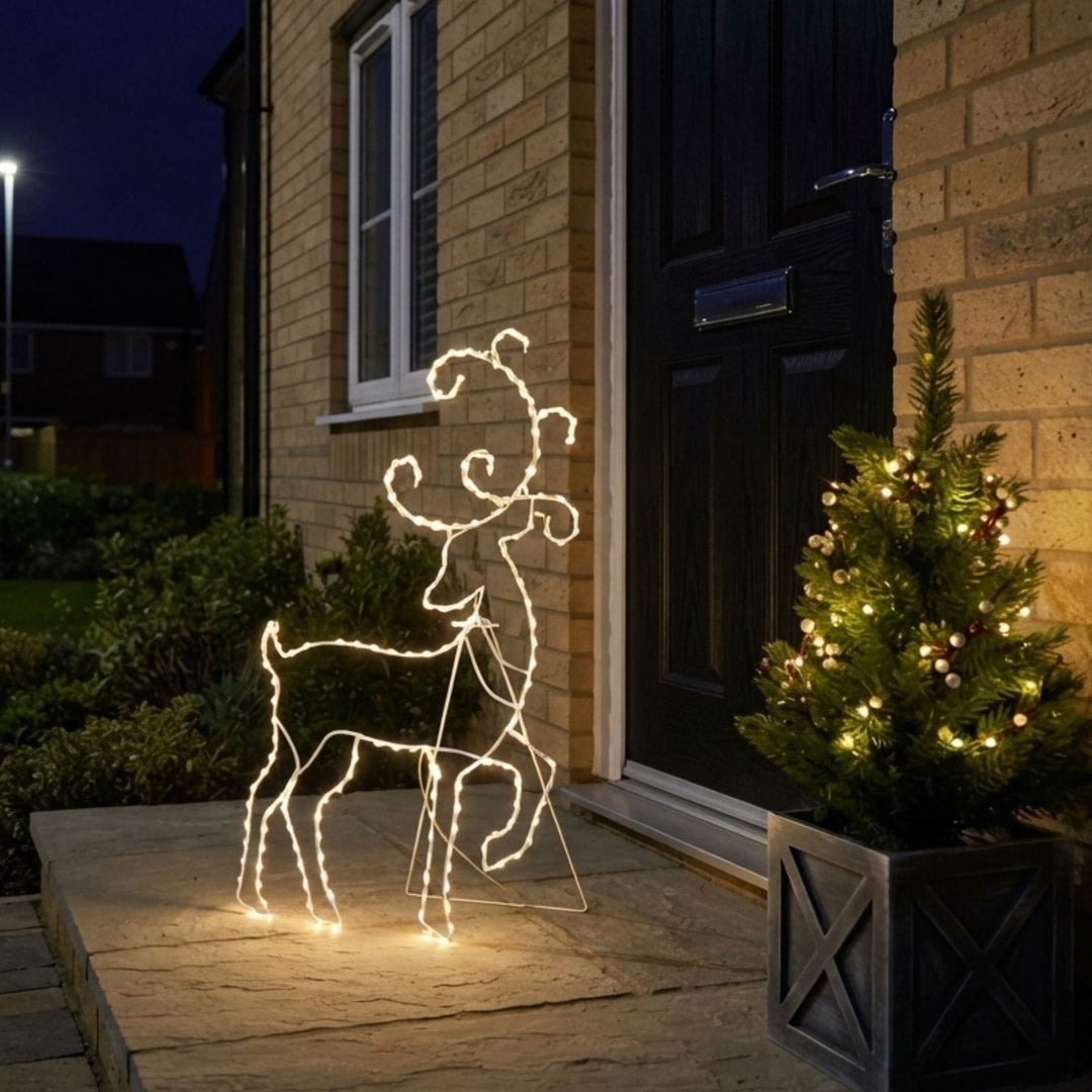A lighted deer outline holiday decoration standing on a stone doorstep at dusk, with a lit Christmas tree and a brick house in the background.