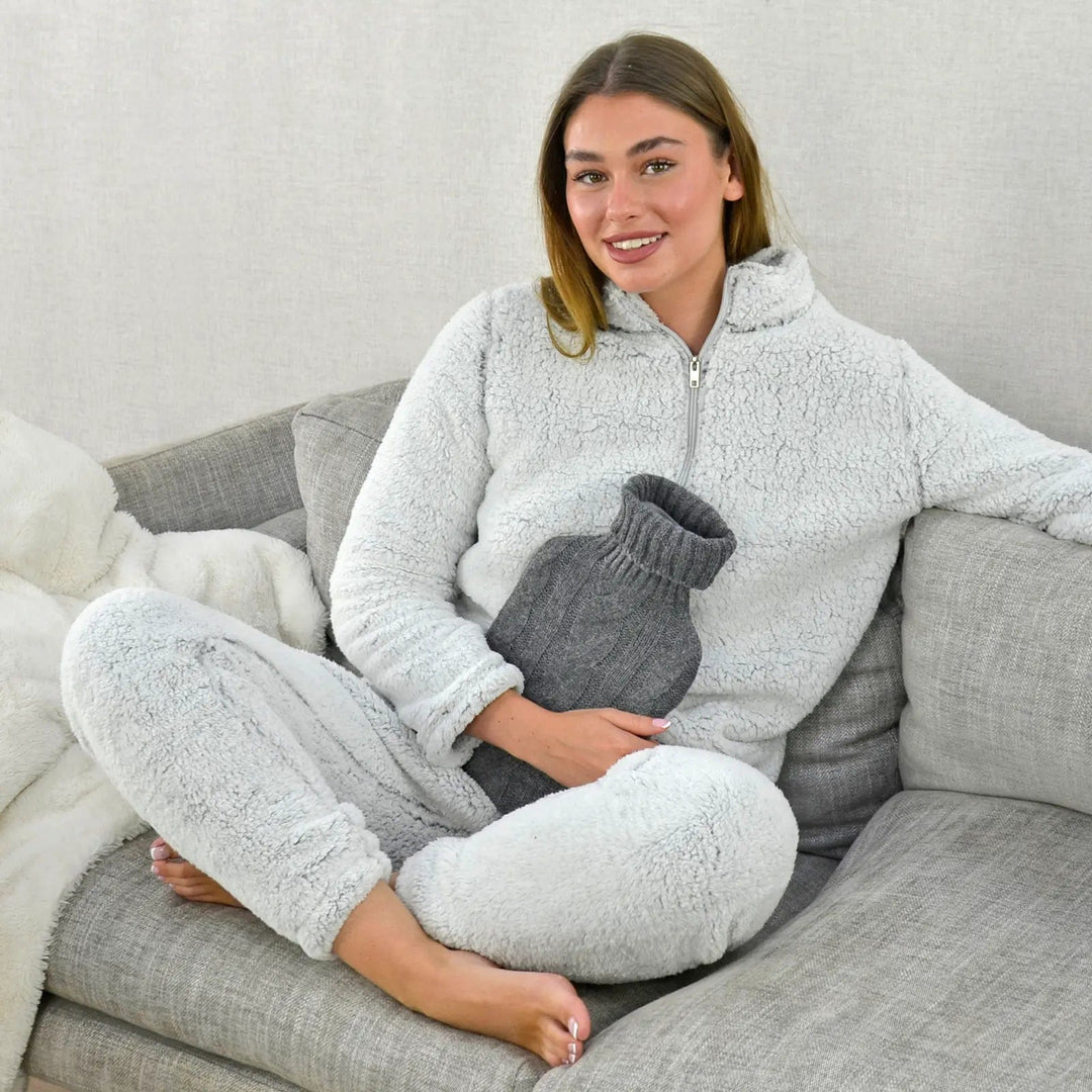 Woman sitting crossed legged holding a hot water bottle with chunky grey cable knit cover