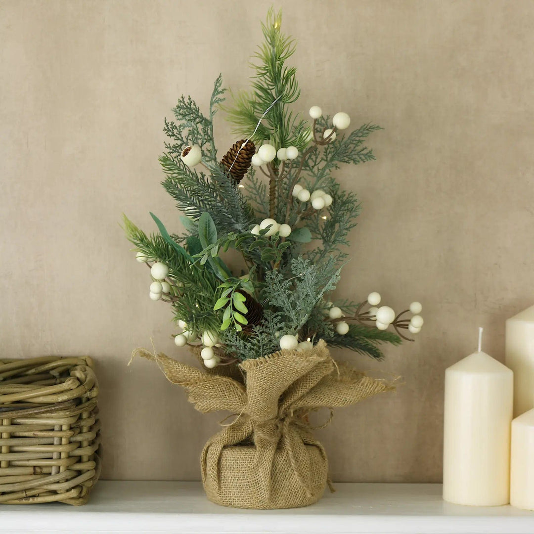 Christmas tree with lights, white berries and pine cones, with jute base on a shelf with white candles