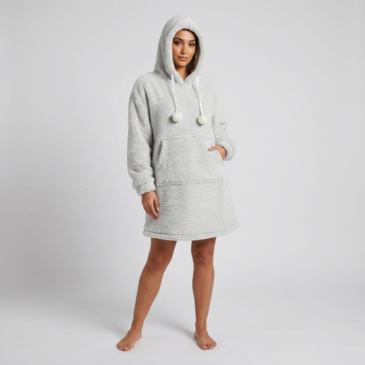 A woman modeling a light gray heathered sherpa hoodie dress with white drawstrings and pom-poms, standing against a light gray studio background.