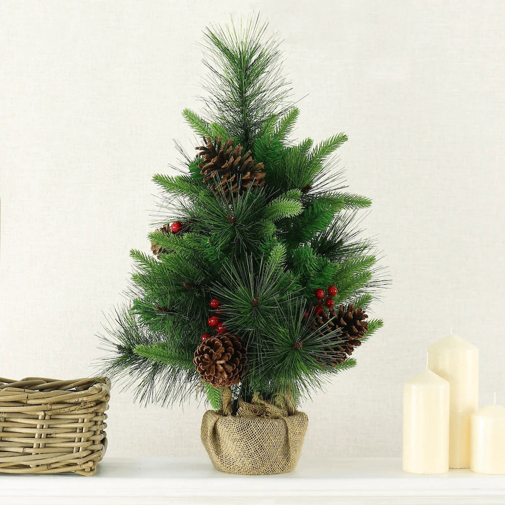 Small green pine Christmas tree with cones, berries and a hessian wrapped base on a table with candles