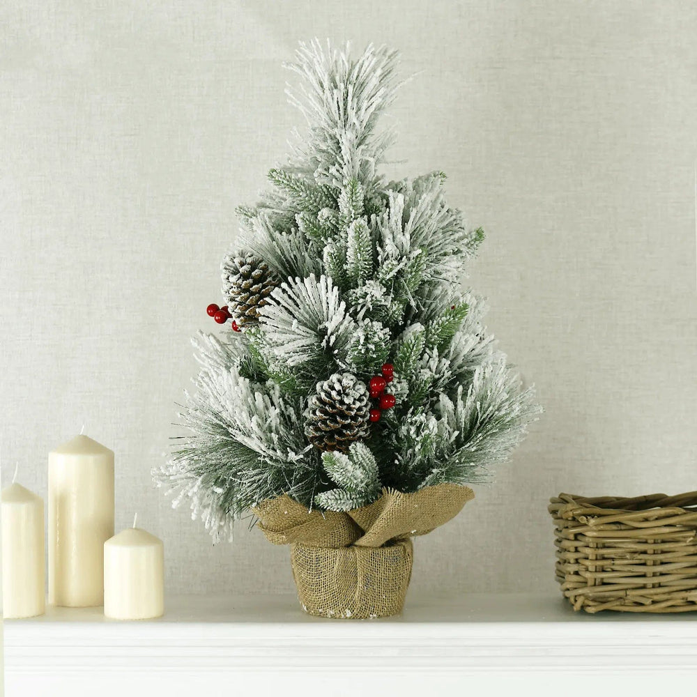 Small Christmas tree with snowy branches, pine cones, berries and hessian covered base on a table with cream coloured candles