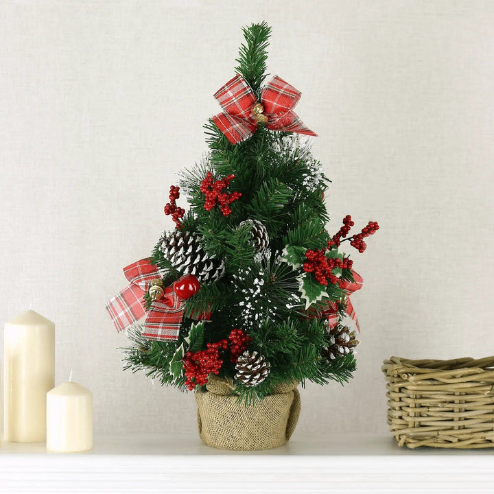 Small pre decorated Christmas tree with red tartan ribbons, pine cones and berries on a table with candles