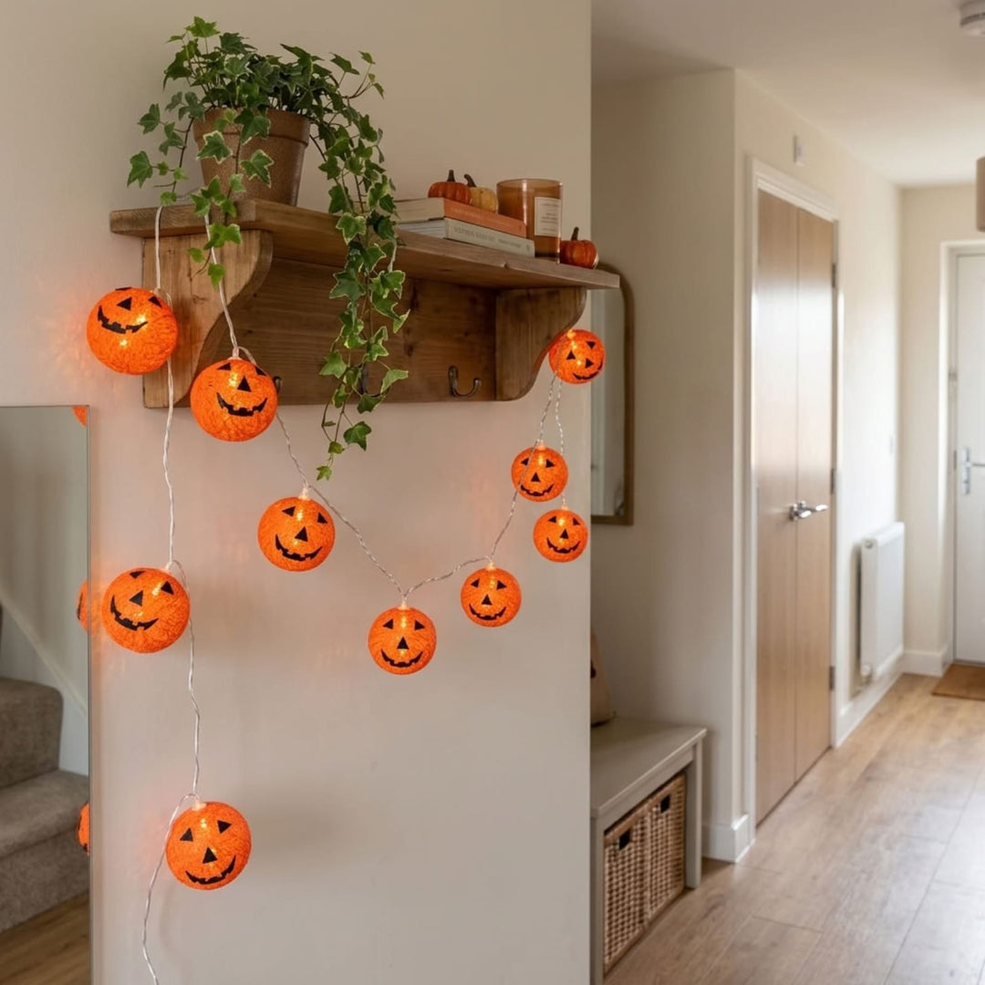 Orange jack-o'-lantern string lights hanging in a curved garland on a white wall in a home hallway, with a wooden shelf and green trailing plant above.