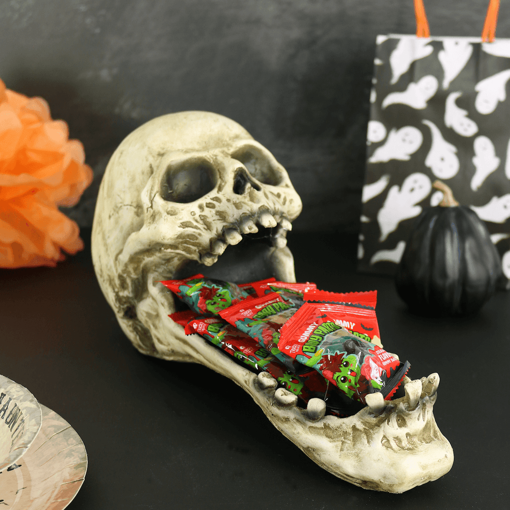 latex skeleton halloween treat tray on black table holding a variety of gummy halloween sweets with black pumpkin, ghost treat bag and halloween paper plates in the background