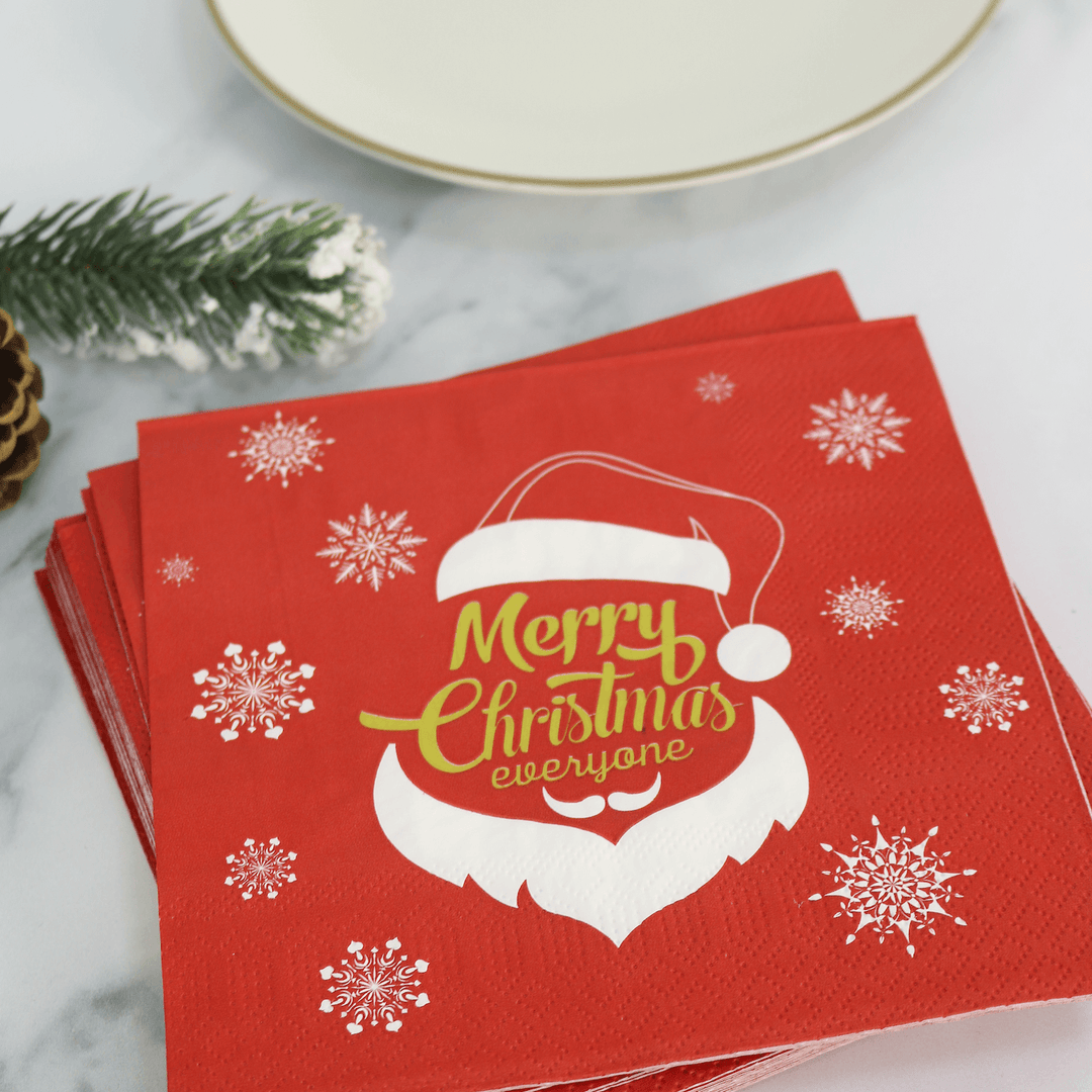 a pile of red christmas merry christmas napkins on a table with plate and pine cone decorations in the background