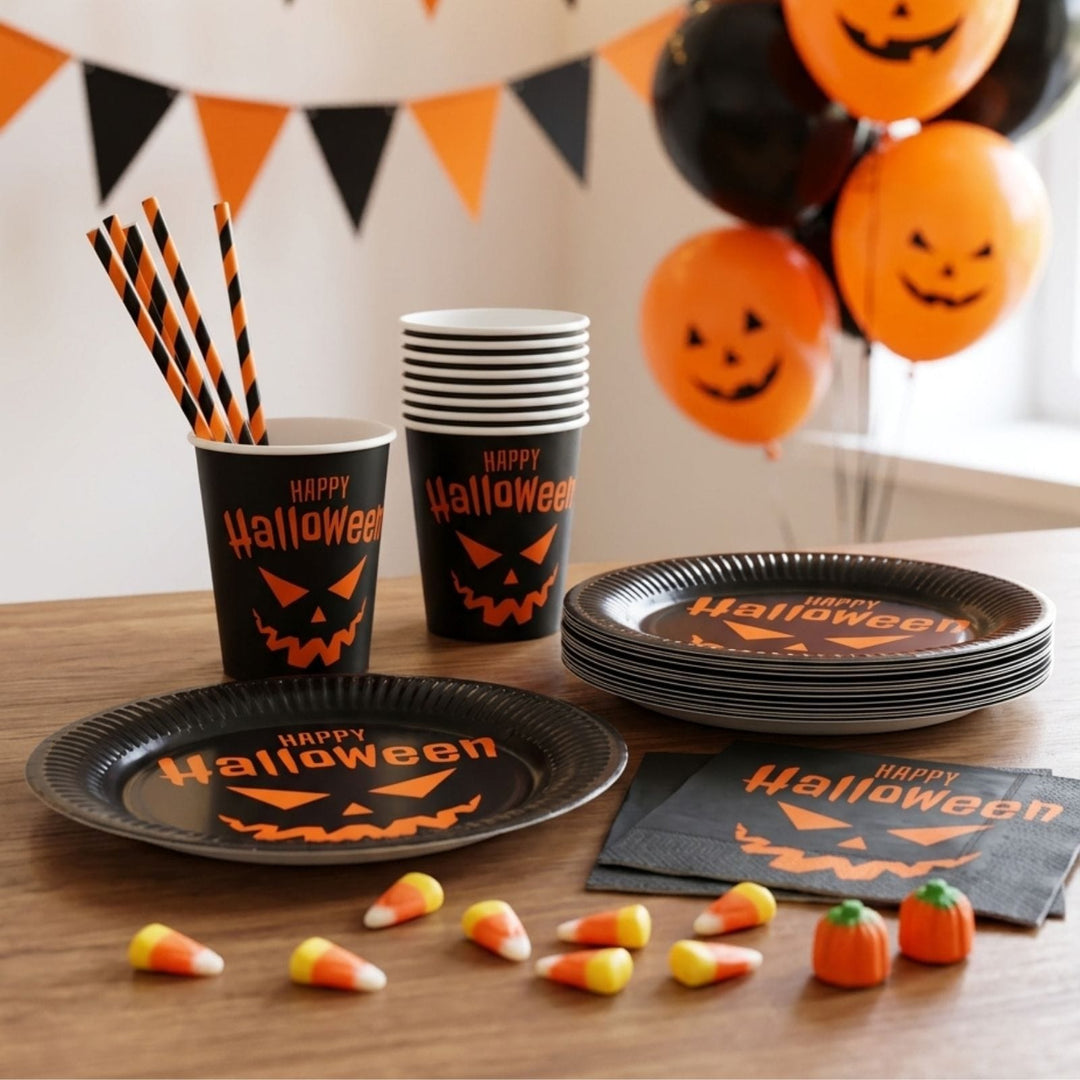 Halloween party tableware set including black paper plates, cups, straws, and napkins with orange 'Happy Halloween' text and jack-o'-lantern faces, arranged on a wooden table with candy corn and pumpkin-shaped balloons in the background.
