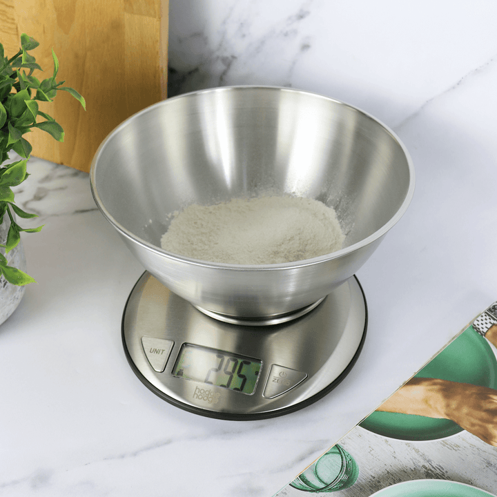 kitchen scale on marble worktop beside recipe book, chopping board and plant weighing flour in stainless steel bowl