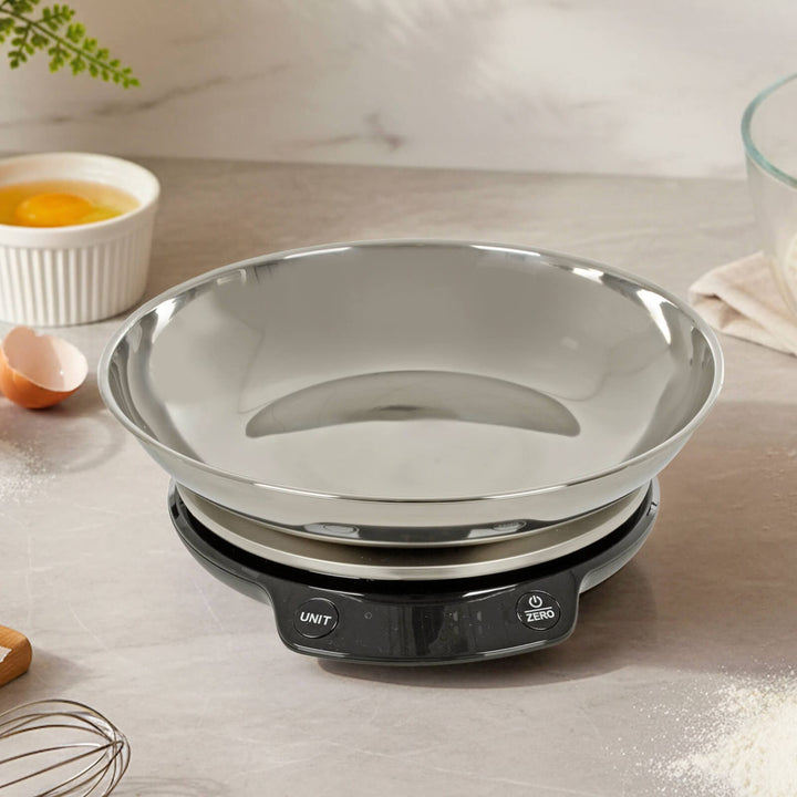 Stainless steel kitchen scales on a worktop with eggs, flour and a whisk
