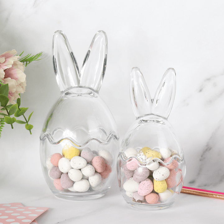 large and medium sizes bunny ears storage jars on marble background filled with mini eggs beside pink flower