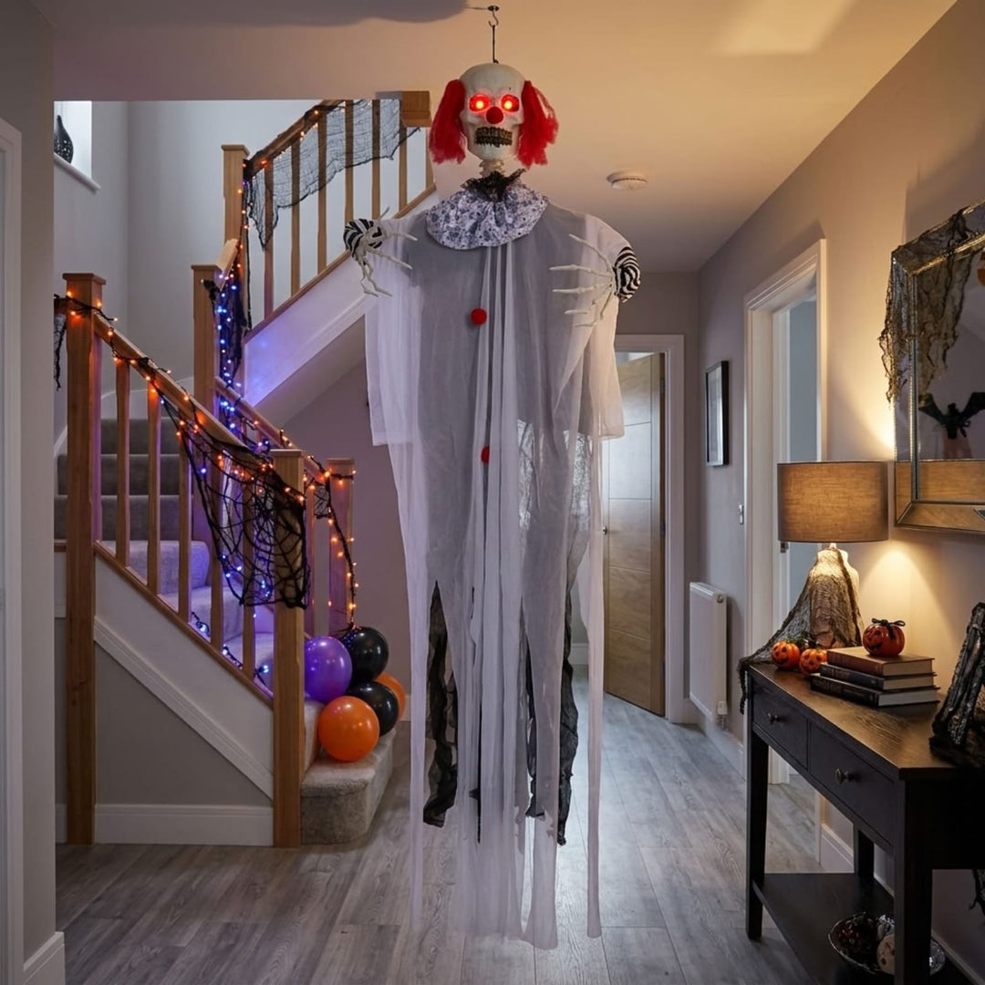 A hanging Halloween decoration shaped like a creepy clown or ghostly figure with red hair, a white tattered robe, and glowing red eyes, displayed in a decorated home hallway next to a staircase.