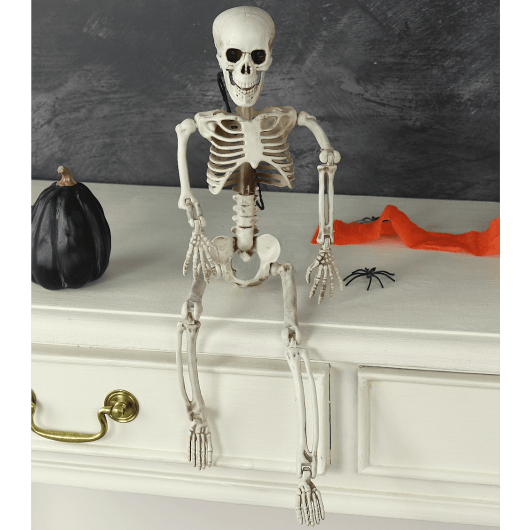 skeleton sitting on white table beside mini black pumpkin and artificial spiders