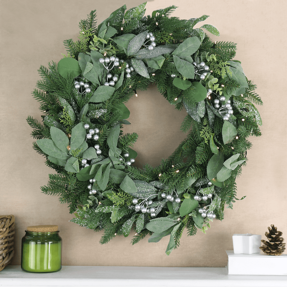 green wreath on beige colour background with fireplace ledge below featuring candles, pine cones and a white book