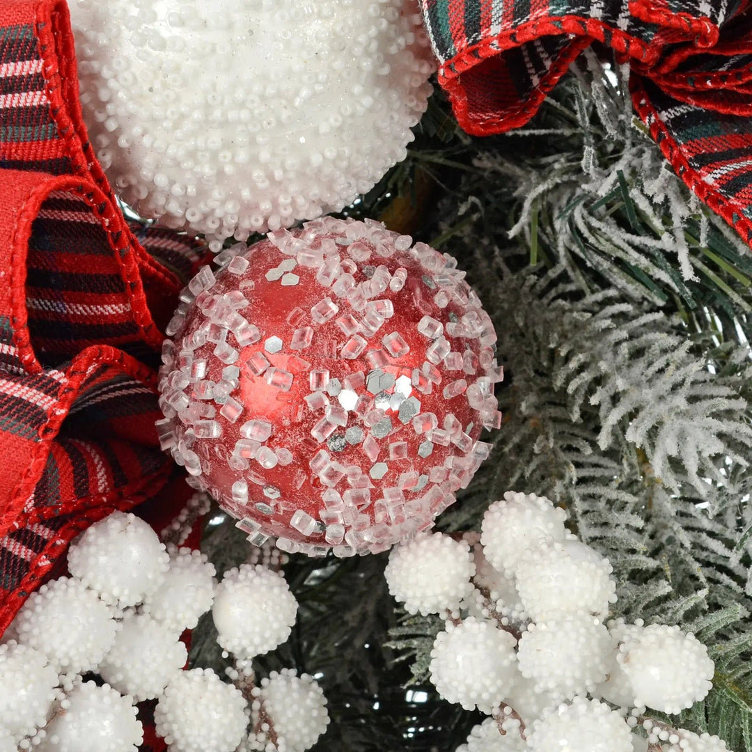 Close up of a red bauble with fake frost pieces, white snow berries, red tartan ribbon and green pine branches