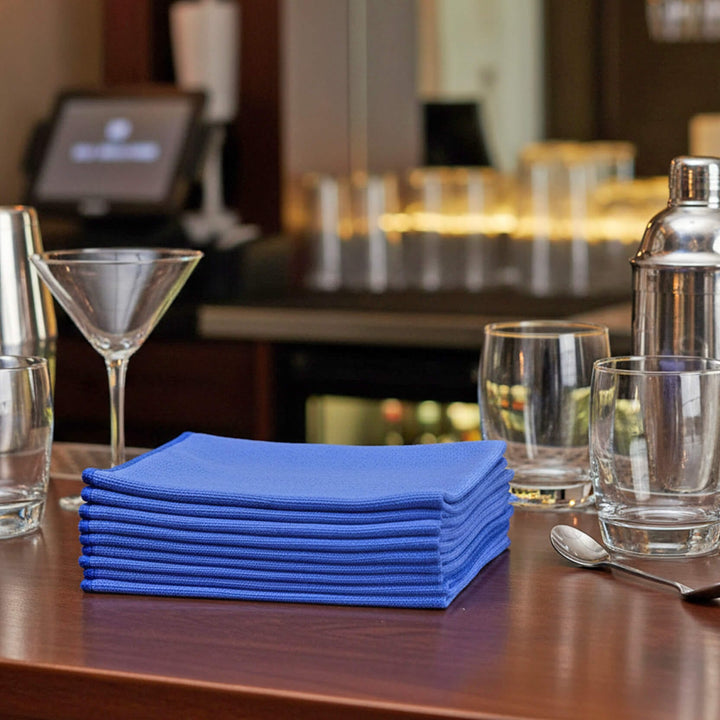 Stack of blue glass cloths on a table with glasses and a cocktail shaker in a blurred bar setting.