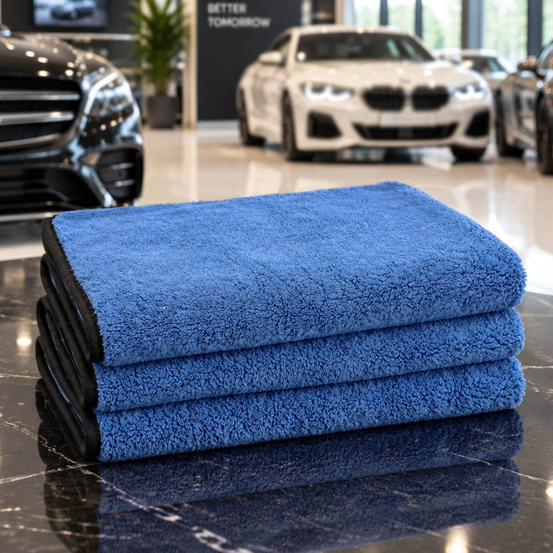 Three folded blue microfiber cleaning towels stacked on a glossy black marble surface in a car showroom, with blurred cars in the background.