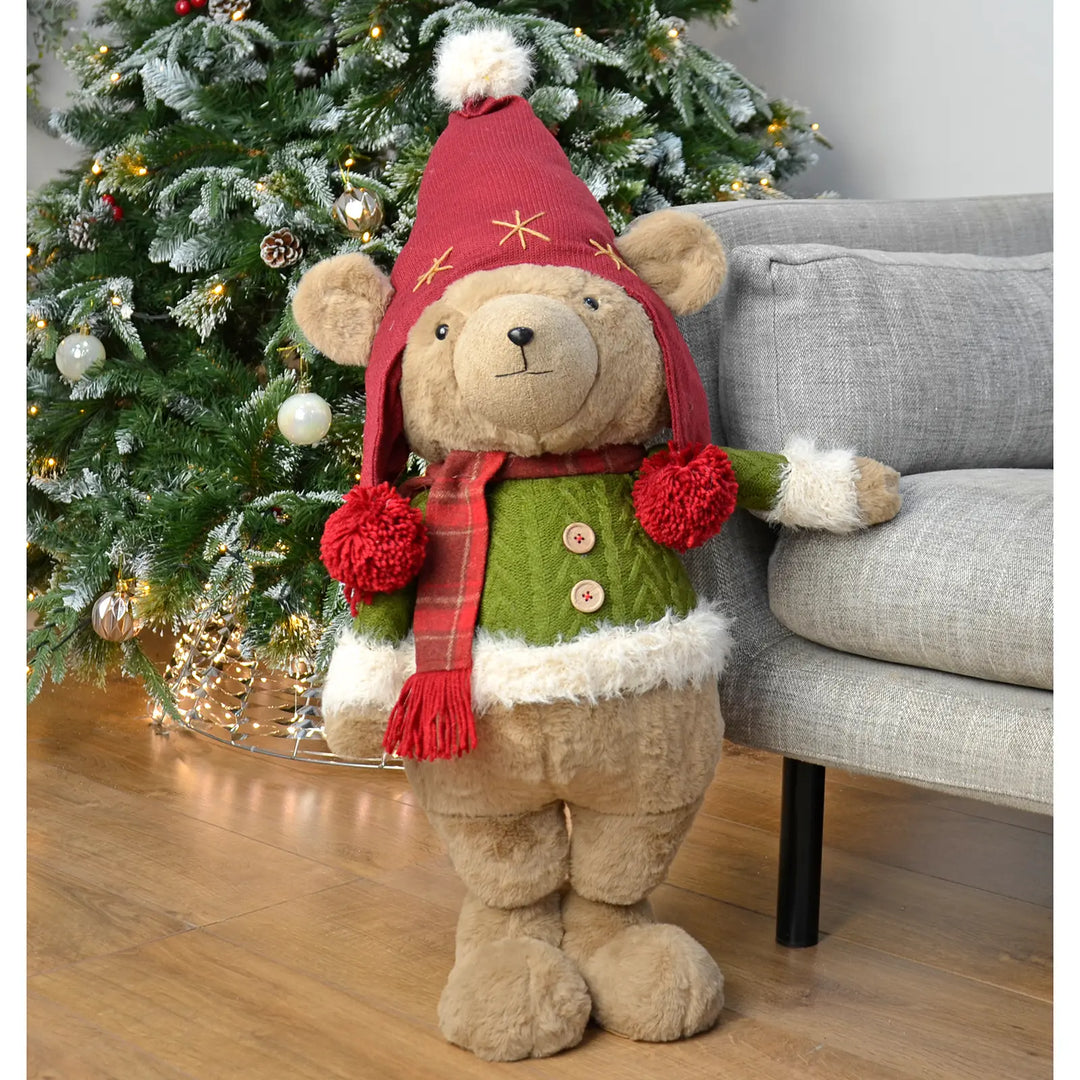 Large standing Christmas bear wearing a green jumper and red trapper hat in front of a decorated tree
