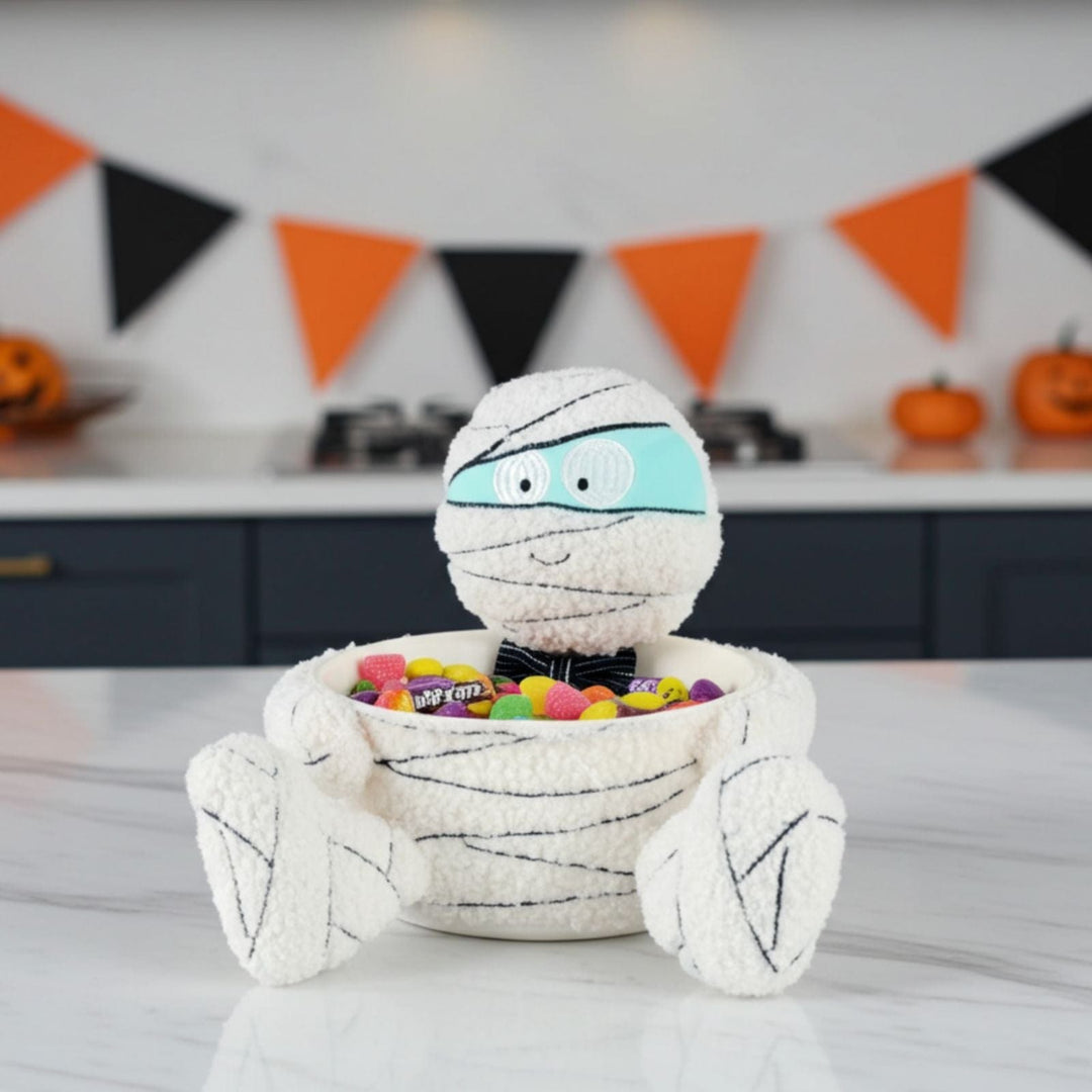 Halloween-themed mummy candy bowl with colorful candies on a table, decorated with Halloween flags in the background.