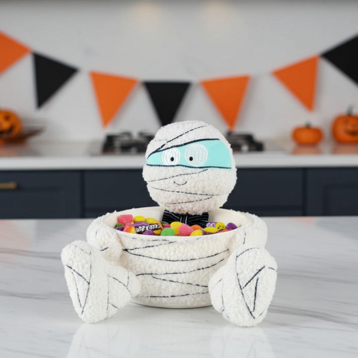 Halloween-themed mummy candy bowl with colorful candies on a table, decorated with Halloween flags in the background.