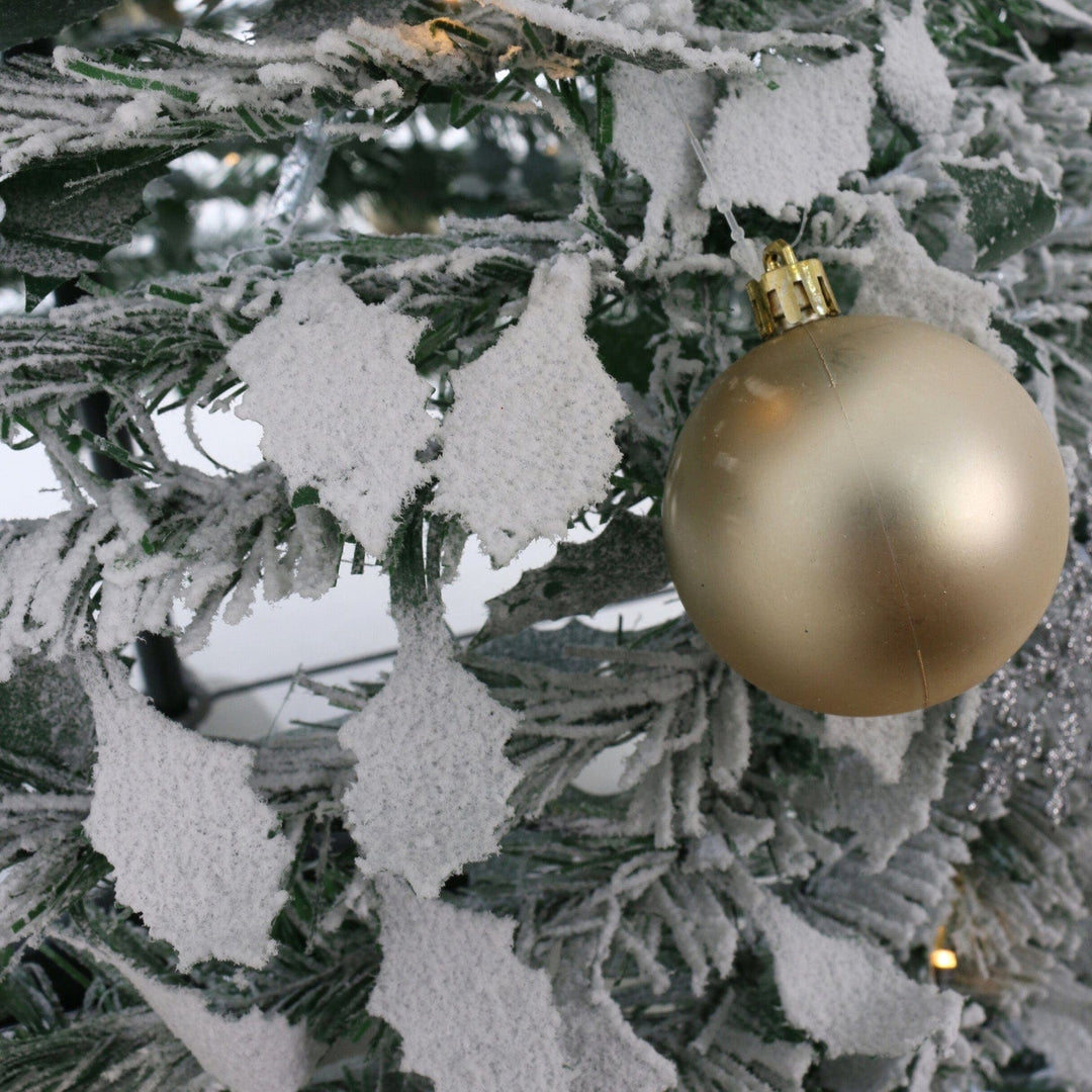 close up of holly leaves flocked with snow beside gold christmas bauble