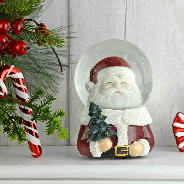 Decorative snow globe with Santa Claus holding a tree, surrounded by Christmas decorations on a white surface.