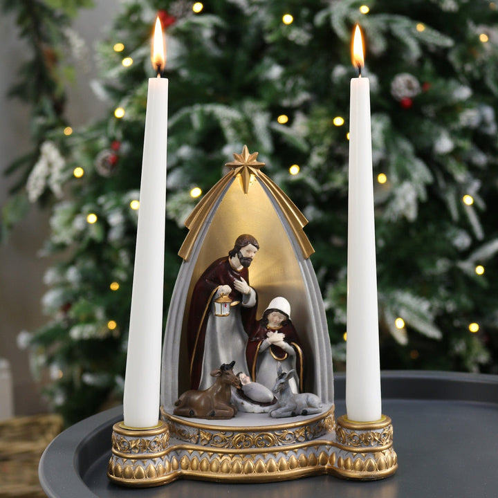 nativity ornament with tapered candles inside and lit on table in front of christmas tree