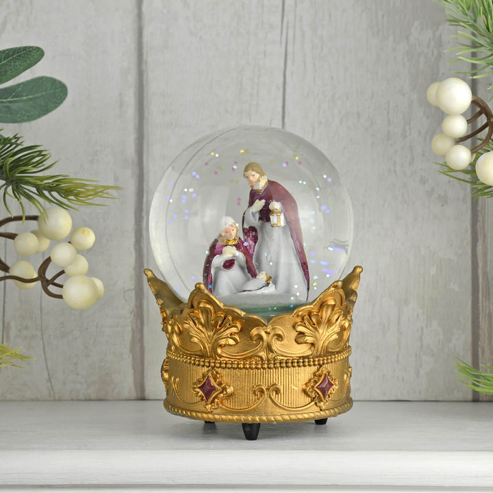 Decorative snow globe with a gold base featuring a Christmas nativity scene, against a light wooden panel background.