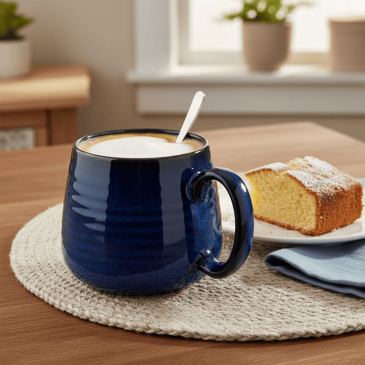 Blue mug with a spoon in a creamy substance on a wooden table with a cake and plants in the background.