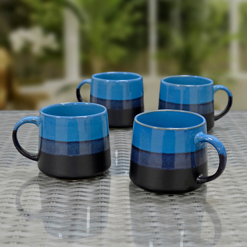 Four blue and black ceramic mugs on a glass topped rattan table with a conservatory background.