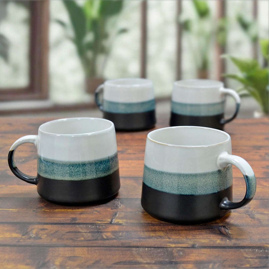 Four large porcelain mugs with white blue and black on a wooden table with plants.