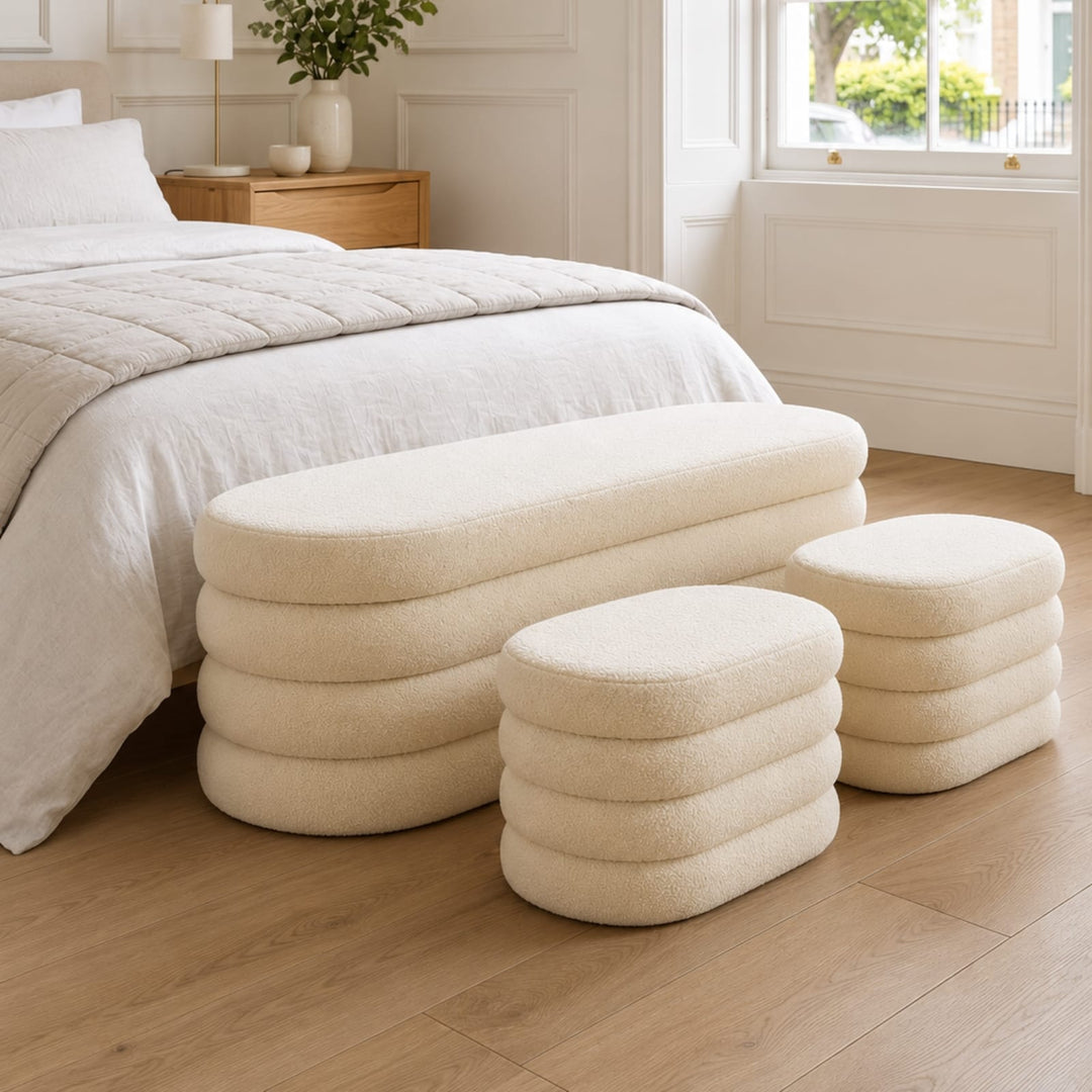 Three cream upholstered ottoman-style bench stools arranged in a bedroom beside a bed with white bedding and light wood flooring.