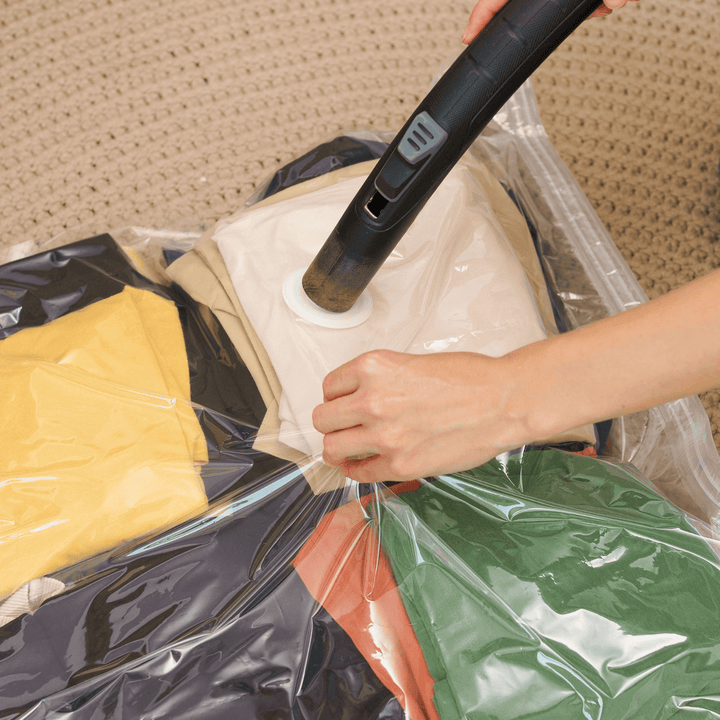 Person using a handheld vacuum cleaner to clean a plastic bag filled with clothes on a carpeted floor.