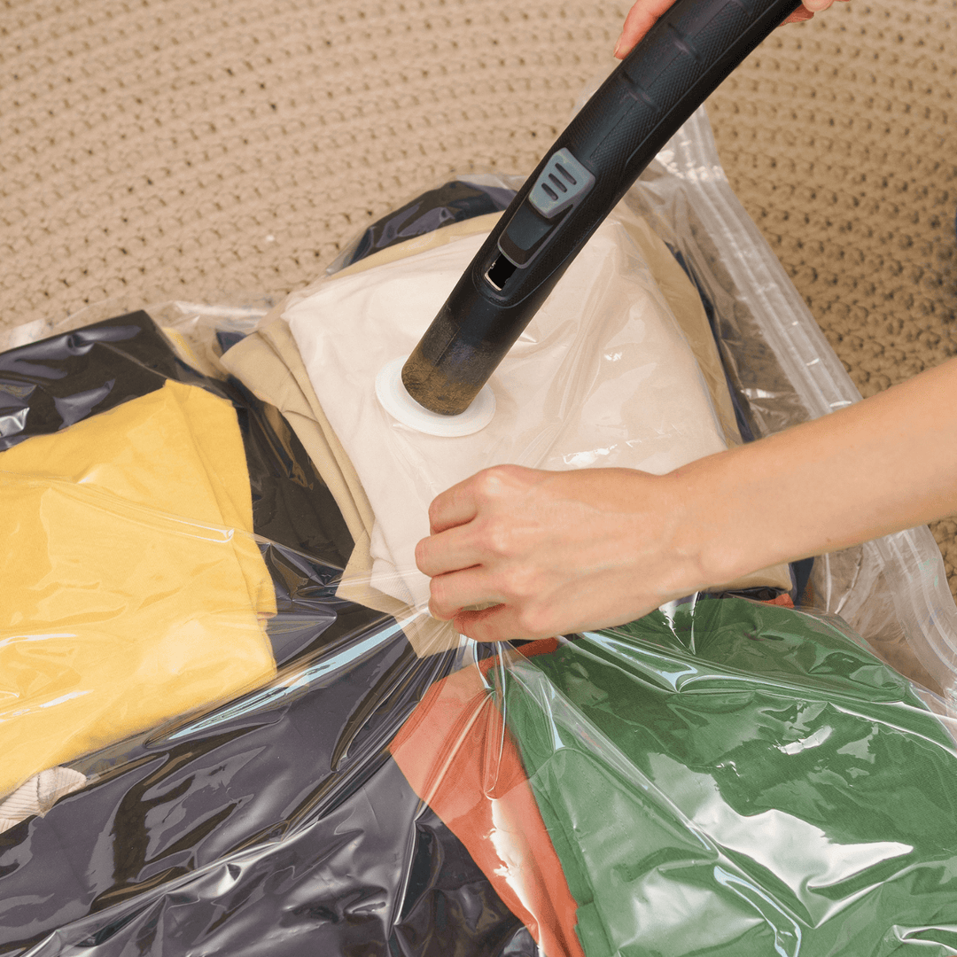 Person using a handheld vacuum cleaner to clean a plastic bag filled with clothes on a carpeted floor.