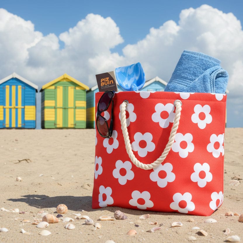 Red beach bag with white flower pattern and rope handles on a beach with brightly coloured beach huts and blue sky